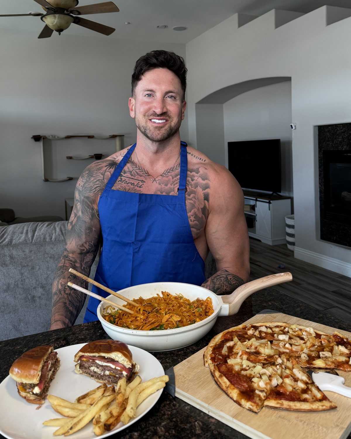Troy Adashun in his kitchen with a burger and fries, a pizza and a bowl of pasta from the book